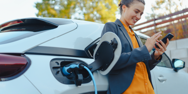 Mulher sorrindo, usando celular enquanto carro recarrega carro elétrico. 